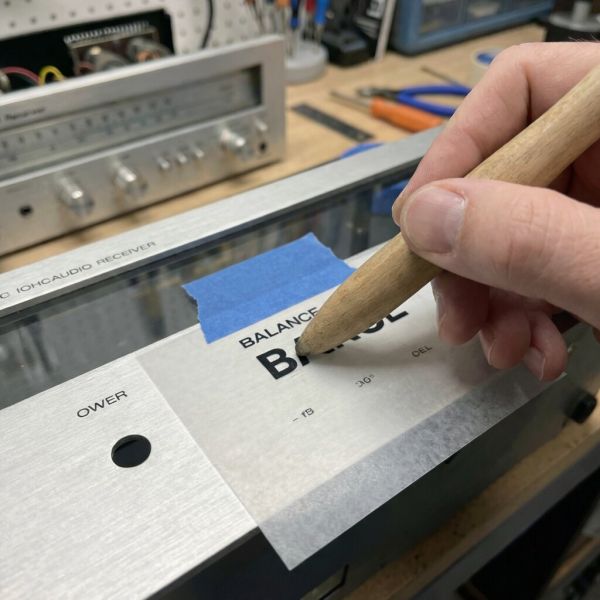 A person using a wooden stylus to burnish dry transfer lettering onto a vintage audio receiver faceplate.
